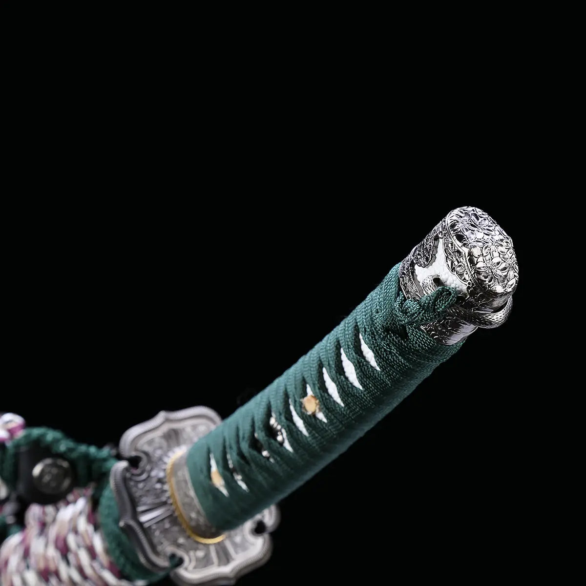 Détail du manche du katana japonais artisanal Seiryū no Tachi photographié sur fond noir uni. Le tsuka est recouvert d’un tsuka-ito vert foncé tressé serré, laissant apparaître une same blanche en losanges. Le pommeau (kashira) et la garde (tsuba) en métal sculpté sont visibles, avec des finitions argentées et un assemblage traditionnel japonais.
