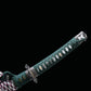 Détail rapproché du manche du katana japonais artisanal Seiryū no Tachi photographié sur fond noir uni. Le tsuka est habillé d’un tsuka-ito vert foncé tressé, laissant apparaître une same blanche en losanges réguliers. Les menuki métalliques décoratifs sont visibles sous le tressage, ainsi que la garde traditionnelle en métal sculpté et le sageo multicolore attaché à la base du manche.
