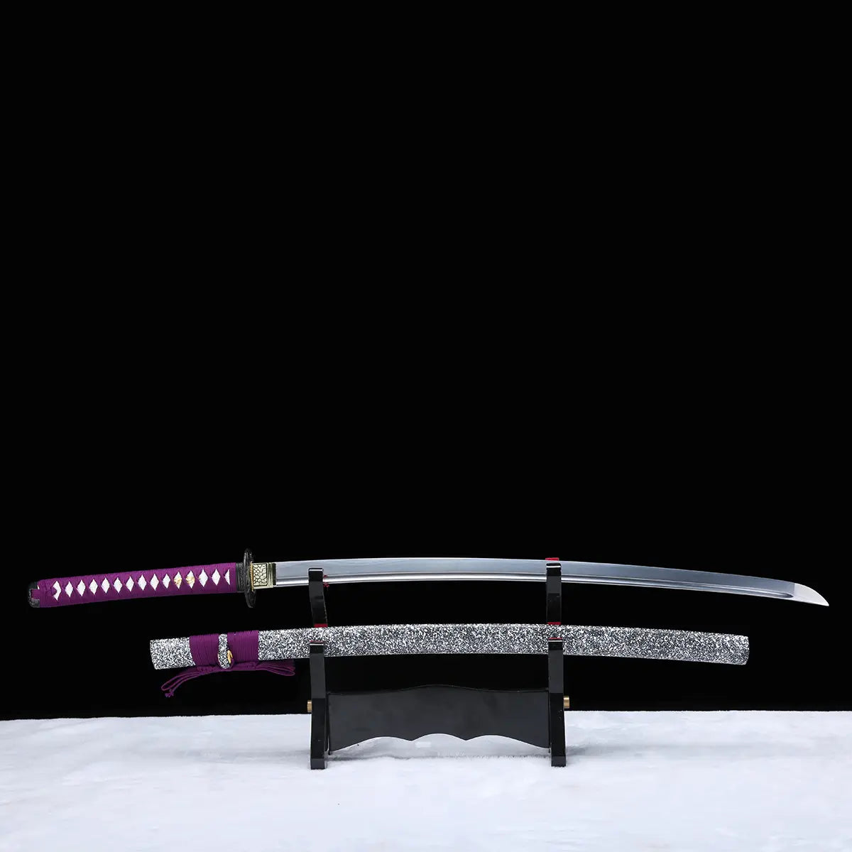 Katana japonais artisanal Murasaki no Kage exposé sur un support en bois avec la lame dégainée au-dessus du fourreau, photographié sur fond noir avec surface blanche, poignée violette tressée avec peau de raie blanche, tsuba noire, lame courbe polie et saya noir laqué à éclaboussures blanches avec cordon sageo violet.

