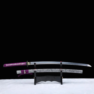 Katana japonais artisanal Murasaki no Kage exposé sur un support en bois avec la lame dégainée au-dessus du fourreau, photographié sur fond noir avec surface blanche, poignée violette tressée avec peau de raie blanche, tsuba noire, lame courbe polie et saya noir laqué à éclaboussures blanches avec cordon sageo violet.
