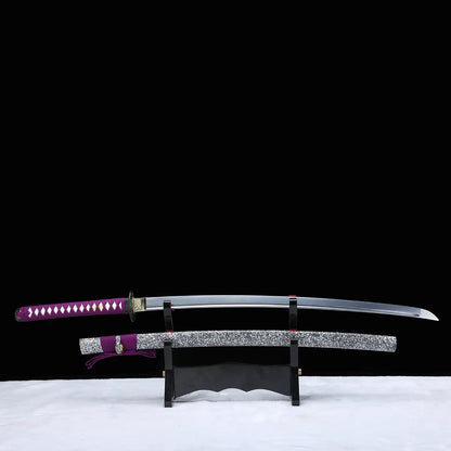 Katana japonais artisanal Murasaki no Kage exposé sur un support en bois avec la lame dégainée au-dessus du fourreau, photographié sur fond noir avec surface blanche, poignée violette tressée avec peau de raie blanche, tsuba noire, lame courbe polie et saya noir laqué à éclaboussures blanches avec cordon sageo violet.
