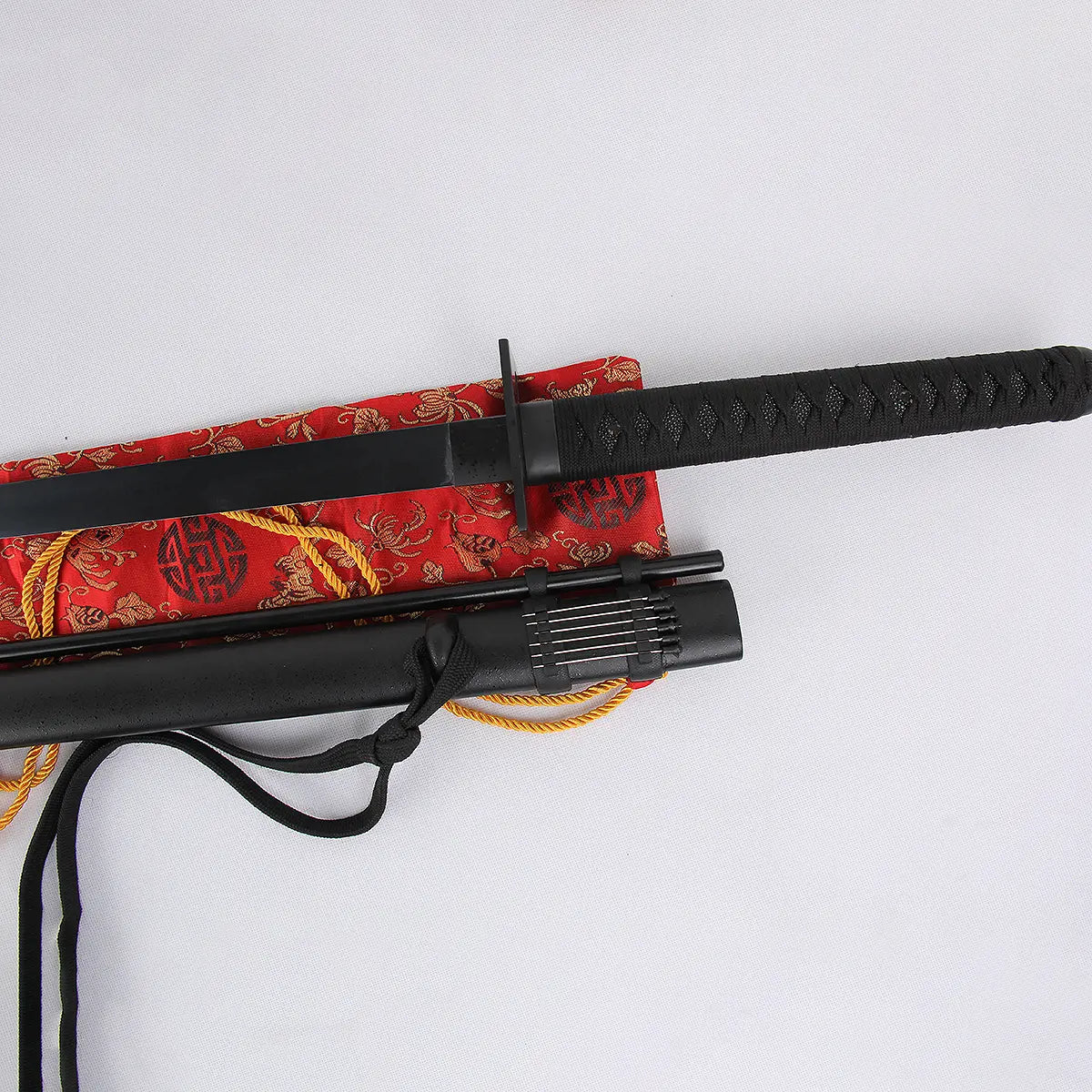 Vue rapprochée du katana japonais artisanal “Shinobi” sur fond blanc : lame noire partiellement dégainée, tsuba carrée, tsuka noire tressée, fourreau noir mat avec porte-aiguilles décoratif et petit tube, posé sur sac en brocart rouge à cordon jaune — katana ninja, ninjatō, katana noir, Blade Bushido