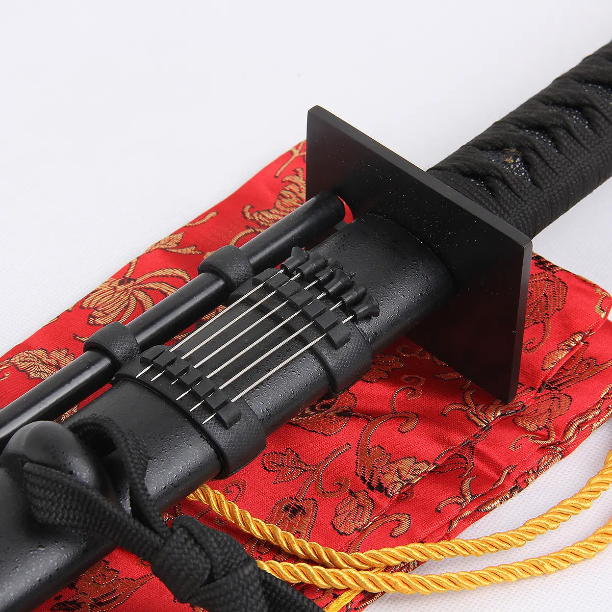 Gros plan près de la garde du katana “Shinobi” : tsuba carrée noire, tsuka tressée noire, saya noir mat avec tube décoratif et porte-aiguilles alignant 12 aiguilles inox, posé sur sac en brocart rouge à cordelette dorée — katana ninja, ninjatō, détail monture, Blade Bushido.