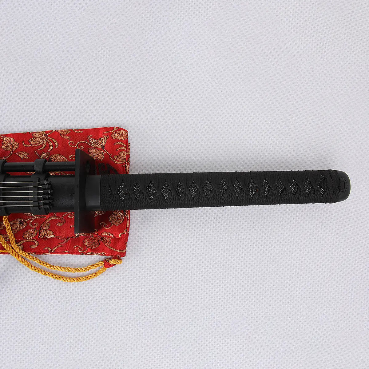 Poignée tsuka noire tressée du katana “Shinobi” photographiée à l’horizontale sur fond blanc, garde tsuba carrée visible, extrémité du saya noir mat et porte-aiguilles en arrière-plan, posé partiellement sur sac en brocart rouge à cordelette dorée — katana ninja, ninjatō, détail manche, Blade Bushido.