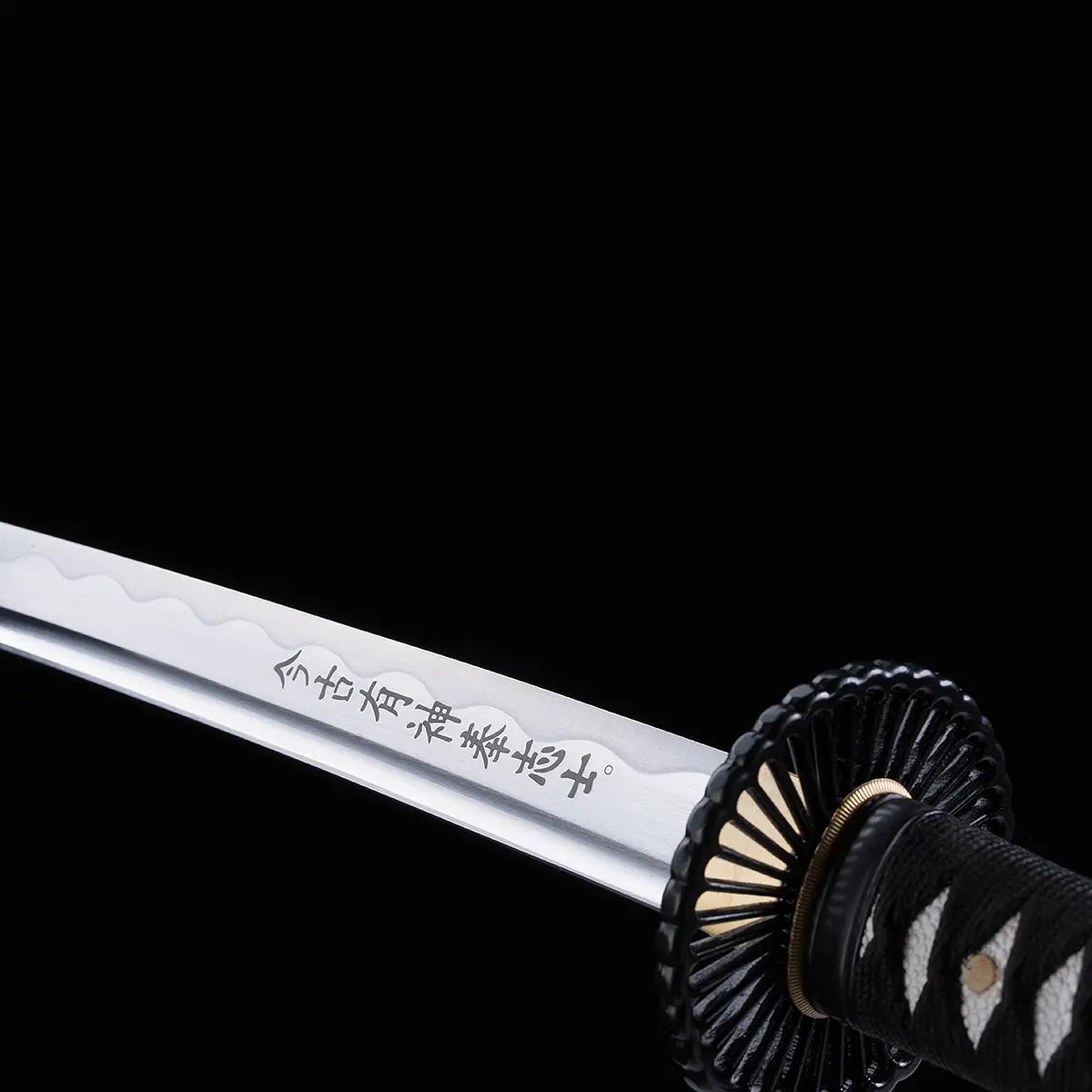 Gros plan sur la lame gravée du katana japonais artisanal Shinshi no Ken sur fond noir profond, acier poli avec hamon ondulé visible, inscription japonaise nette le long de la lame, tsuba noire nervurée et manche noir avec tressage traditionnel laissant apparaître un samegawa blanc.
