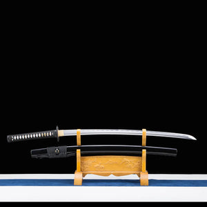 Vue complète du katana japonais artisanal Shinshi no Ken lame dégainée sur un support en bois sculpté doré, fond noir profond avec base claire et tissu bleu, lame en acier poli avec hamon ondulé visible, fourreau noir laqué posé en dessous avec sageo gris foncé, poignée noire avec tressage traditionnel et samegawa blanc apparent.
