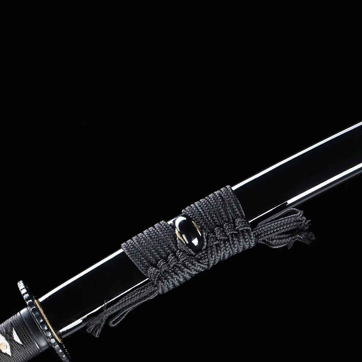 Détail du fourreau (saya) noir laqué du katana japonais artisanal Shinshi no Ken sur fond noir, sageo gris foncé soigneusement noué autour du kurikata, surface brillante et lisse reflétant la lumière, tsuba noire visible à l’avant et manche partiellement apparent.
