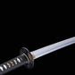 Détail de la lame du katana japonais artisanal Shinshi no Ken sur fond noir profond, acier poli avec ligne de hamon visible, habaki doré à la base de la lame, tsuba noire nervurée, manche noir avec tressage traditionnel laissant apparaître un samegawa blanc et des menuki dorés.
