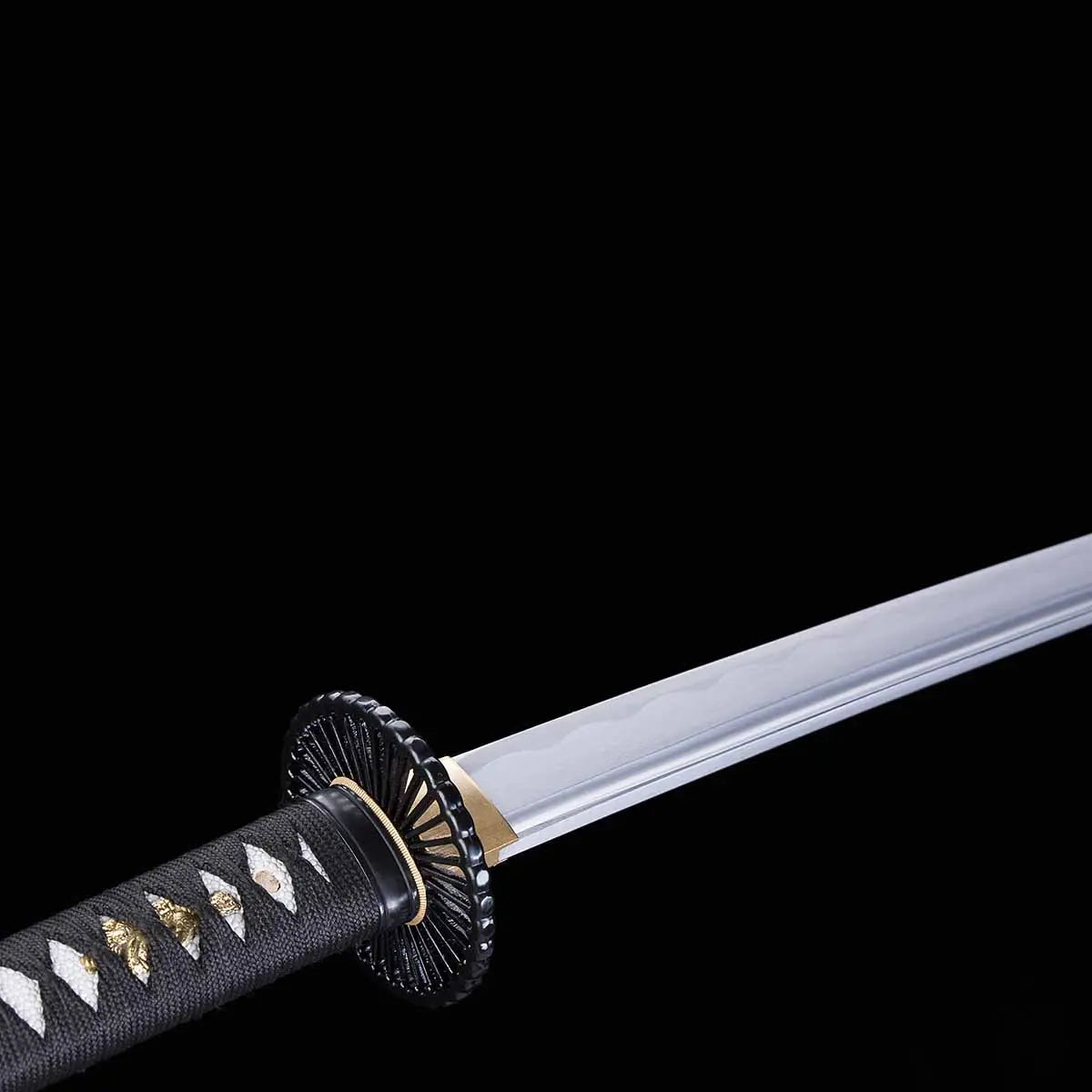 Détail de la lame du katana japonais artisanal Shinshi no Ken sur fond noir profond, acier poli avec ligne de hamon visible, habaki doré à la base de la lame, tsuba noire nervurée, manche noir avec tressage traditionnel laissant apparaître un samegawa blanc et des menuki dorés.
