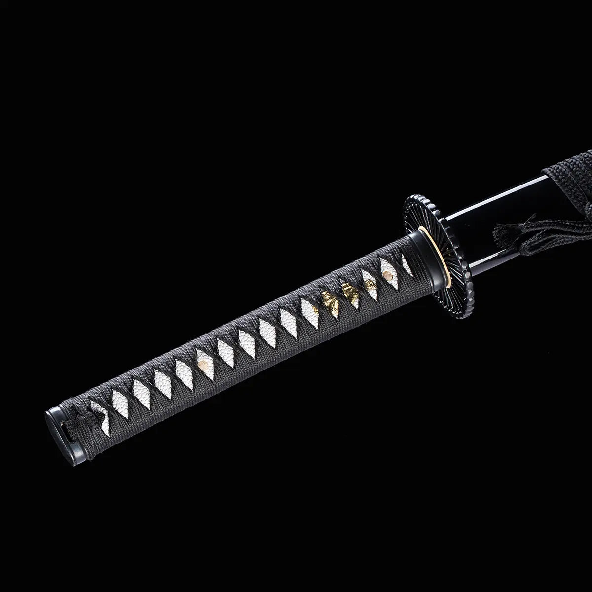Détail du manche (tsuka) du katana japonais artisanal Shinshi no Ken sur fond noir profond, poignée noire avec tressage en losanges laissant apparaître un samegawa blanc, menuki dorés visibles sous le laçage, tsuba noire nervurée et fourreau noir laqué partiellement visible à l’arrière.
