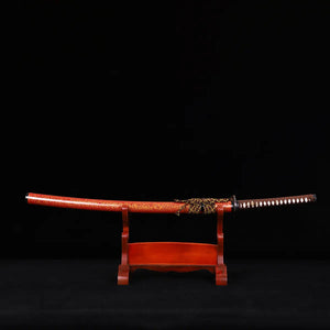 Katana japonais Kannon Ryū au fourreau rouge laqué moucheté or, posé sur un support en bois verni, sur fond noir studio. Sabre artisanal Blade Bushido à hamon authentique en acier 1045 trempé à l’argile.