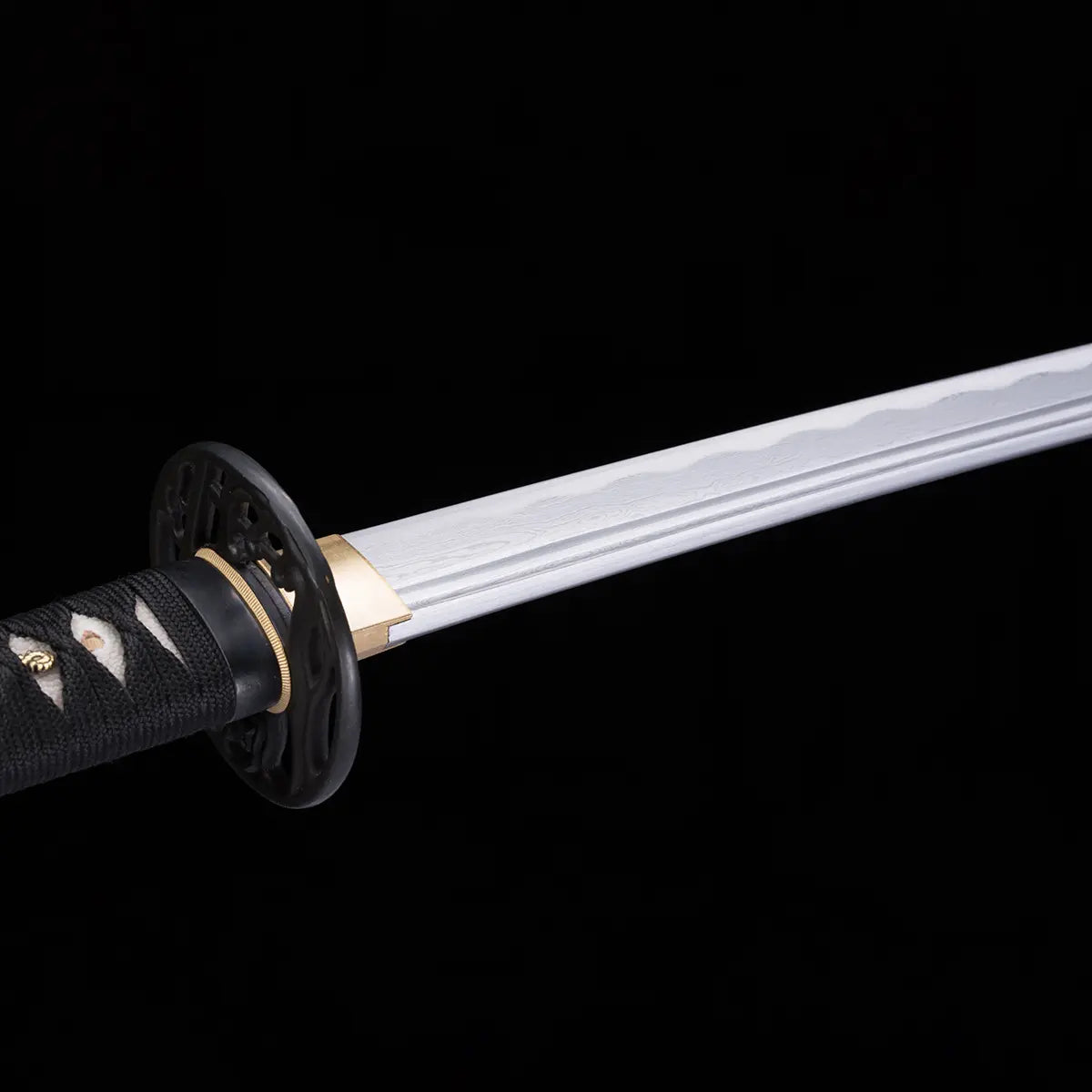 Gros plan sur la lame du katana japonais “Kurenai no Tsuki” en acier damassé avec plis visibles et gorge bohi, garde tsuba ajourée noire et habillage en peau de raie blanche, photographié sur fond noir studio – katana forgé Blade Bushido.