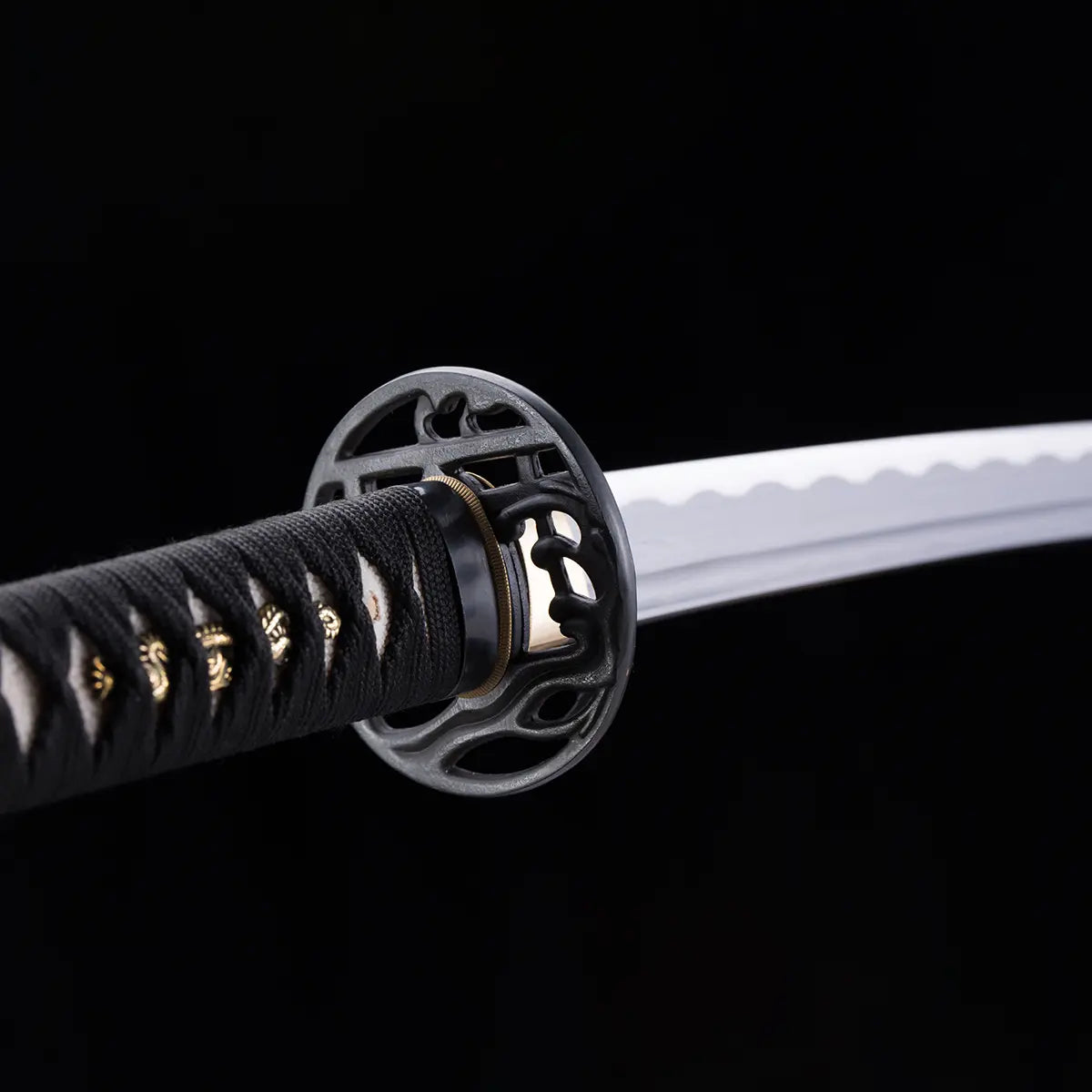 Plan rapproché du katana japonais “Kurenai no Tsuki”, mettant en valeur la garde tsuba ajourée noire, la tsuka noire tressée avec ornements dorés et la lame damassée à hamon ondulé, sur fond noir studio – katana artisanal Blade Bushido.
