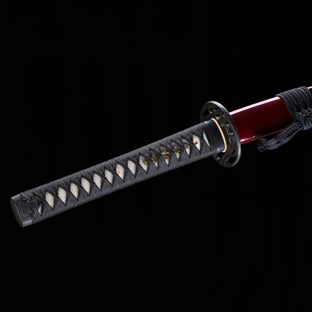 Gros plan du katana japonais “Kurenai no Tsuki”, poignée tsuka noire tressée avec peau de raie blanche, garde tsuba ajourée noire et saya rouge laqué visible sur fond noir studio – katana artisanal Blade Bushido.