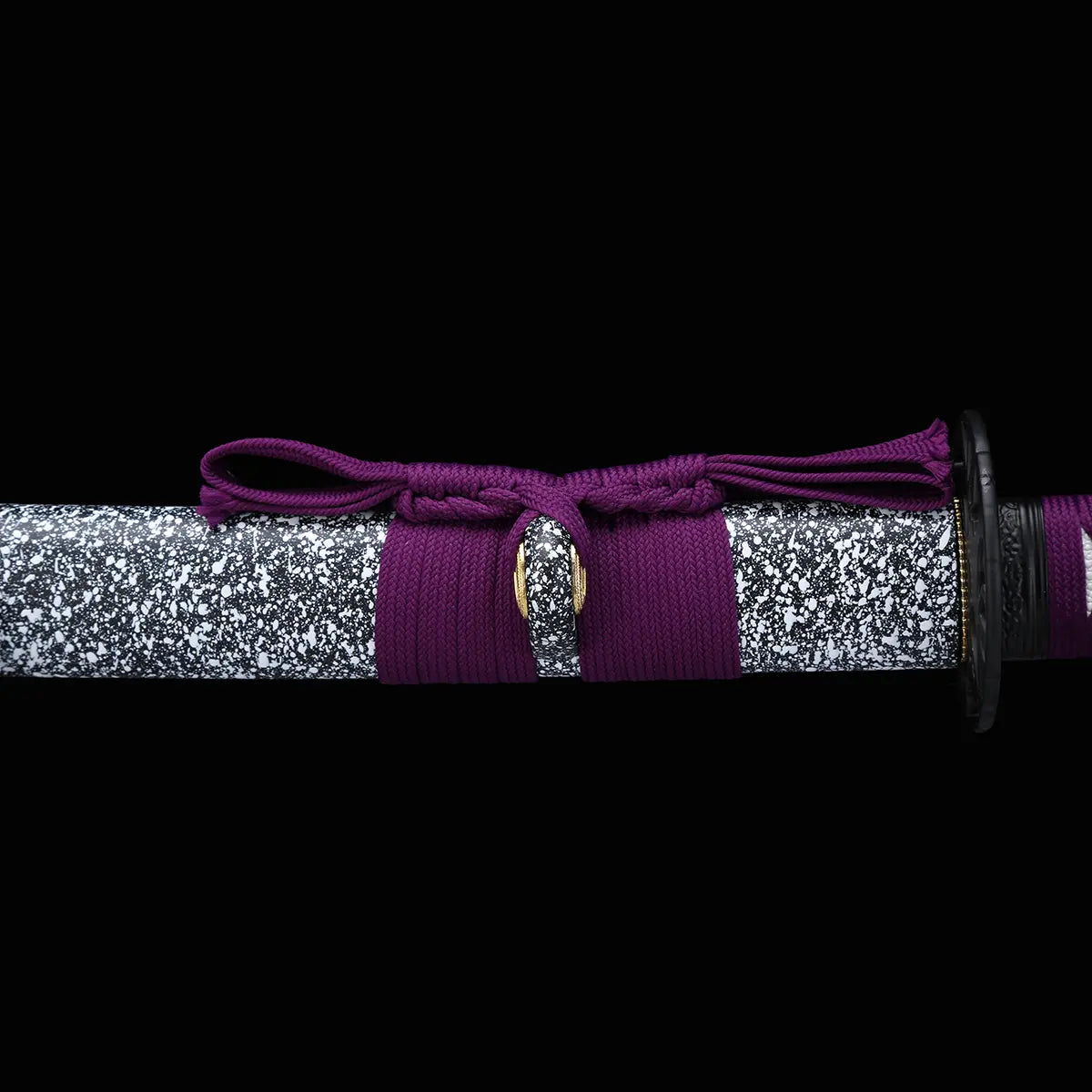 Gros plan du fourreau du katana japonais Murasaki no Kage sur fond noir, saya noir laqué avec motif d’éclaboussures blanches entouré d’un cordon sageo violet noué, avec la tsuba noire partiellement visible à
