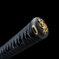 Close-up photograph of the end of a Japanese katana handle on a deep black background, highlighting the black kashira end cap decorated with a gold dragon motif, the black ito wrap tightly braided around the tsuka, and a gold cord ring detail, emphasizing traditional Japanese craftsmanship, premium materials, and refined samurai sword finishing.
