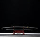 Katana nodachi Monohoshizāo artisanal, lame dégainée posée au-dessus du saya noir laqué sur support en bois (acajou), hamon réel bien visible sur toute la longueur, tsuka noire à losanges ivoire, fond studio noir minimaliste — hommage à Sasaki Kojirō, esprit ōdachi élégant.
