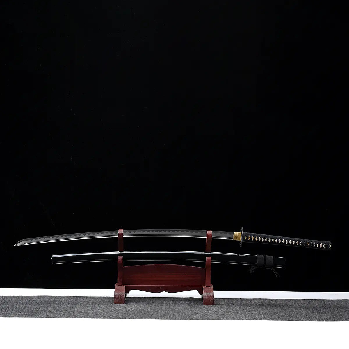 Katana nodachi Monohoshizāo artisanal, lame dégainée posée au-dessus du saya noir laqué sur support en bois (acajou), hamon réel bien visible sur toute la longueur, tsuka noire à losanges ivoire, fond studio noir minimaliste — hommage à Sasaki Kojirō, esprit ōdachi élégant.