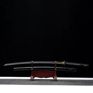 Katana nodachi Monohoshizāo artisanal, lame dégainée posée au-dessus du saya noir laqué sur support en bois (acajou), hamon réel bien visible sur toute la longueur, tsuka noire à losanges ivoire, fond studio noir minimaliste — hommage à Sasaki Kojirō, esprit ōdachi élégant.