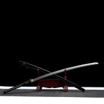 Katana nodachi “Monohoshizāo” artisanal, lame et saya croisés sur support en bois (acajou), hamon réel bien marqué, habaki laiton, tsuka noire à losanges ivoire, fond studio noir minimaliste et tapis gris — hommage à Sasaki Kojirō, esprit ōdachi élancé.