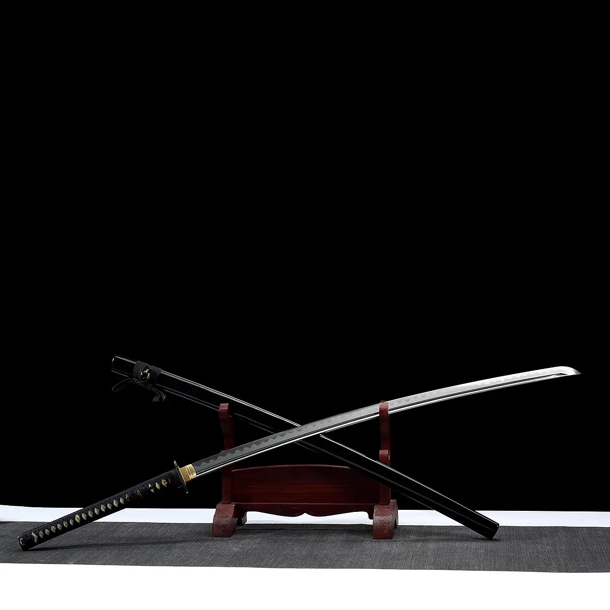 Katana nodachi “Monohoshizāo” artisanal, lame et saya croisés sur support en bois (acajou), hamon réel bien marqué, habaki laiton, tsuka noire à losanges ivoire, fond studio noir minimaliste et tapis gris — hommage à Sasaki Kojirō, esprit ōdachi élancé.