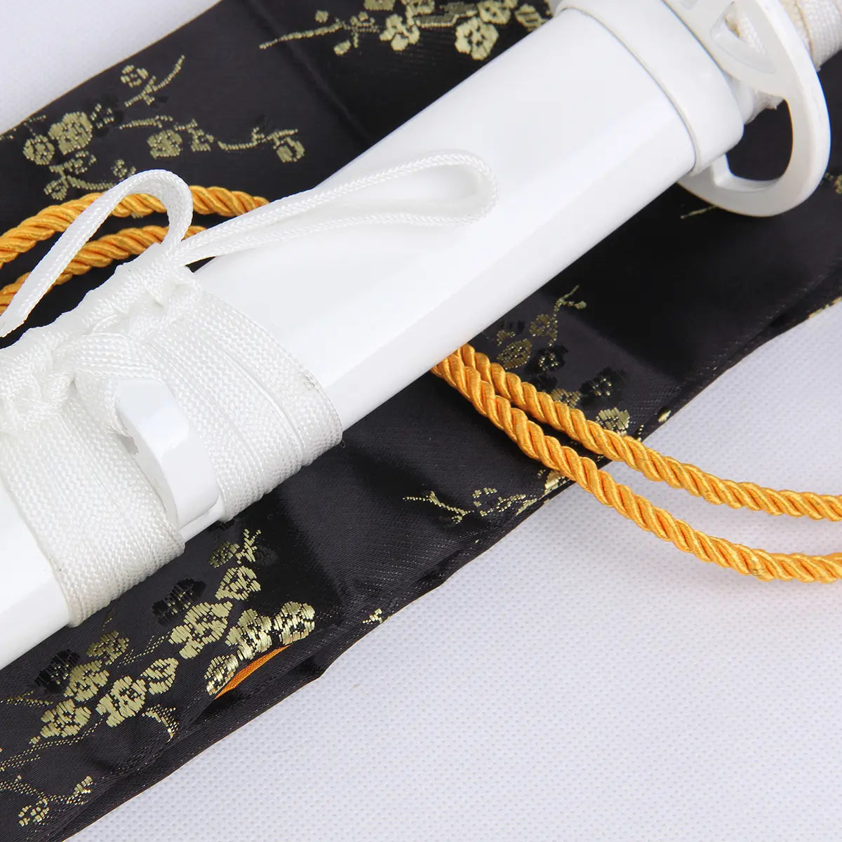 Gros plan du katana Kuchiki Rukia “Sode no Shirayuki” de Bleach, détail du fourreau blanc laqué avec cordon blanc et tressage doré, posé sur une housse noire brodée de motifs floraux dorés.