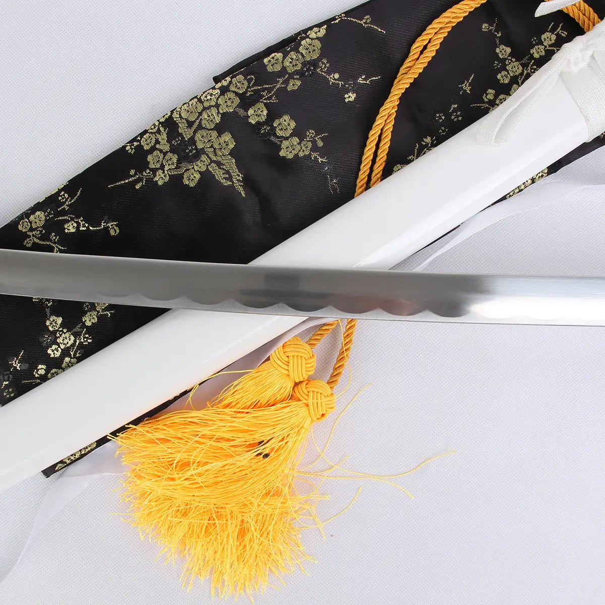 Gros plan du katana Kuchiki Rukia “Sode no Shirayuki” de Bleach, réplique zanpakutō Bankai en acier carbone, montrant la lame brillante au hamon ondulé posée sur le fourreau blanc laqué, avec cordons dorés et housse noire brodée de motifs floraux.