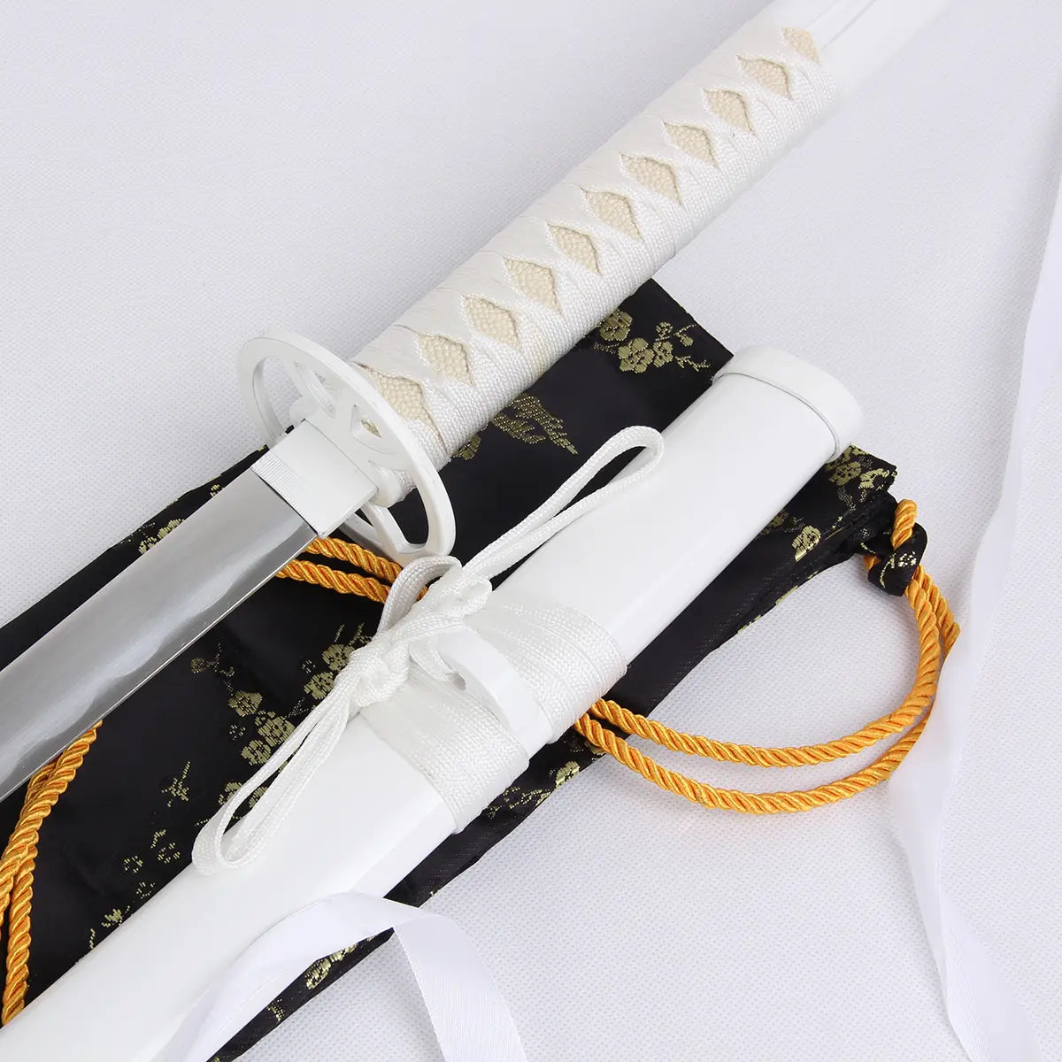 Gros plan du katana Kuchiki Rukia “Sode no Shirayuki” de Bleach, réplique zanpakutō Bankai en acier carbone, montrant la poignée tressée blanche (tsuka) et le fourreau laqué blanc (saya), posés sur une housse noire brodée de motifs dorés avec cordon décoratif jaune.