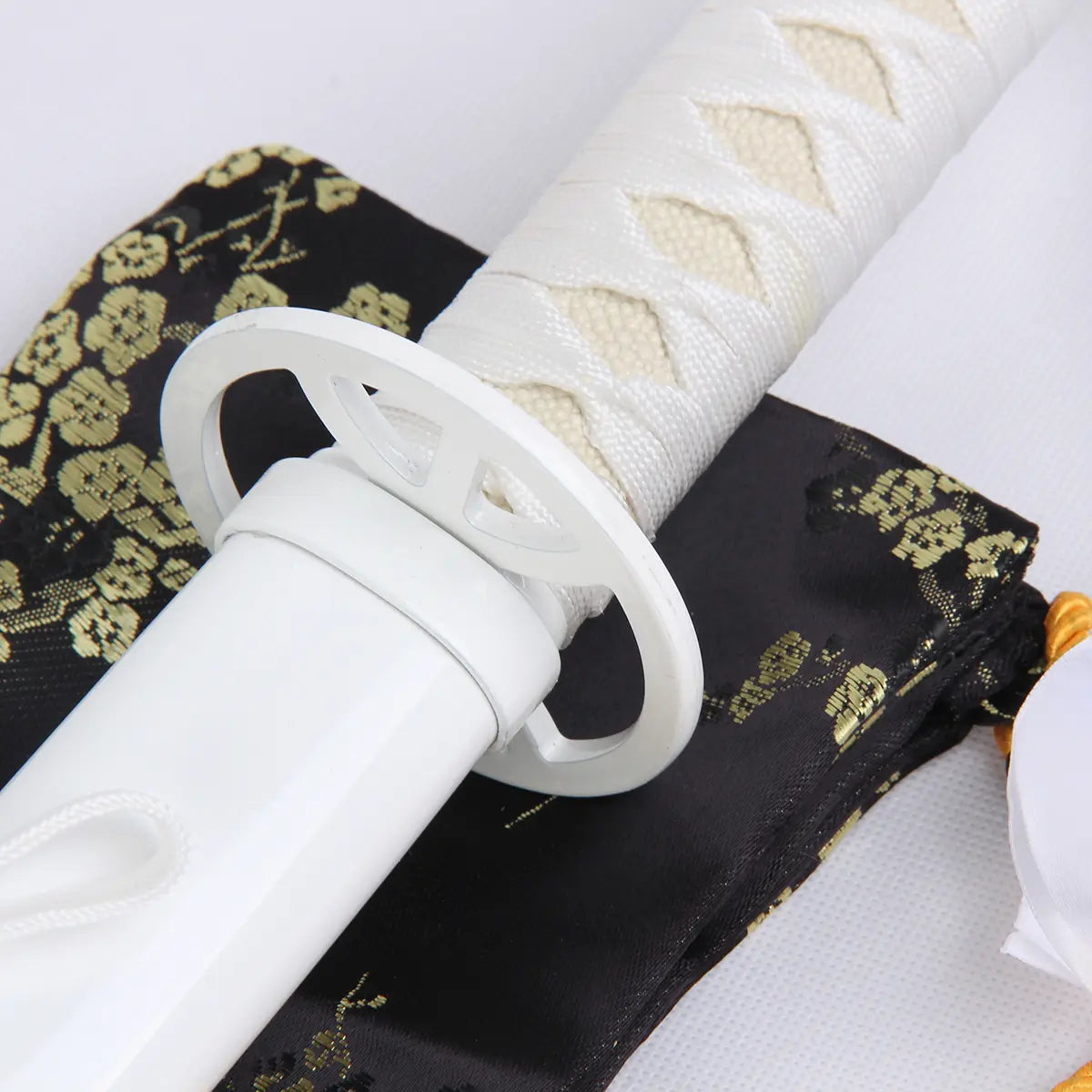 Détail du katana Kuchiki Rukia “Sode no Shirayuki” de Bleach, gros plan sur la garde circulaire blanche (tsuba) et la poignée tressée en blanc, posés sur une housse noire ornée de motifs floraux dorés.
