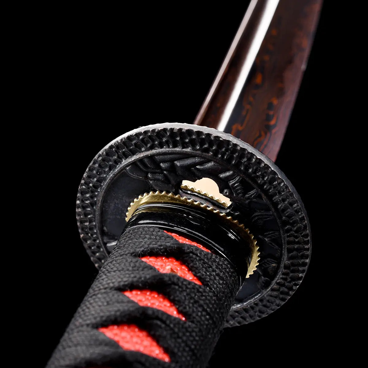 Gros plan sur la garde tsuba du katana japonais Ryū Honō, ornée d’un dragon sculpté en relief, lame damas rouge-feu et poignée tsuka noire tressée de rouge. Fond noir studio Blade Bushido soulignant les détails métalliques et la finition artisanale du sabre.