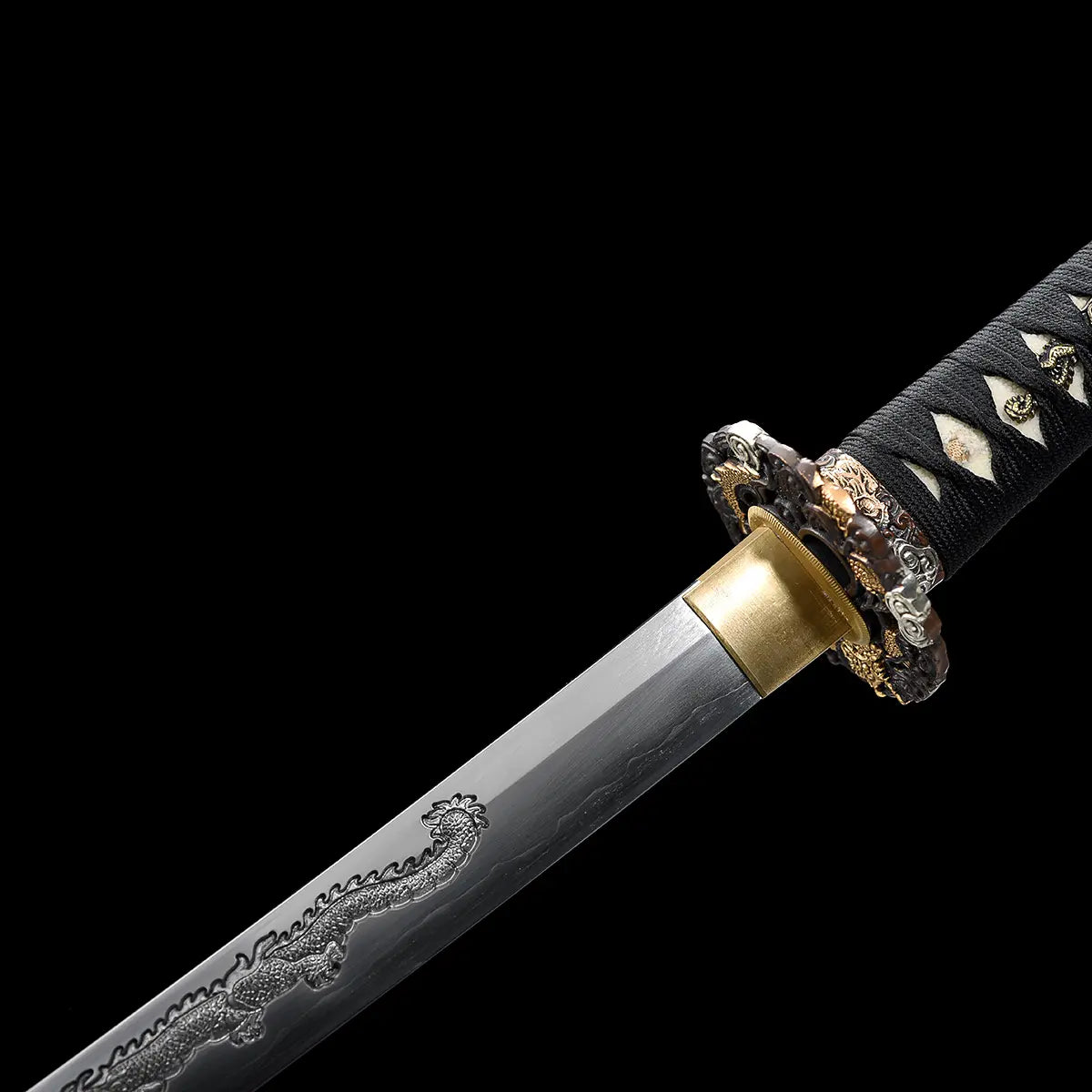 Détail rapproché du katana japonais artisanal Ryū no Ken sur fond noir uniforme, mettant en avant la base de la lame en acier avec gravure dragon finement sculptée, habaki en laiton doré, garde ronde décorée de motifs traditionnels métalliques, manche noir tressé avec losanges blancs en peau de raie, finition nette et contraste marqué entre l’acier poli et les éléments dorés.
