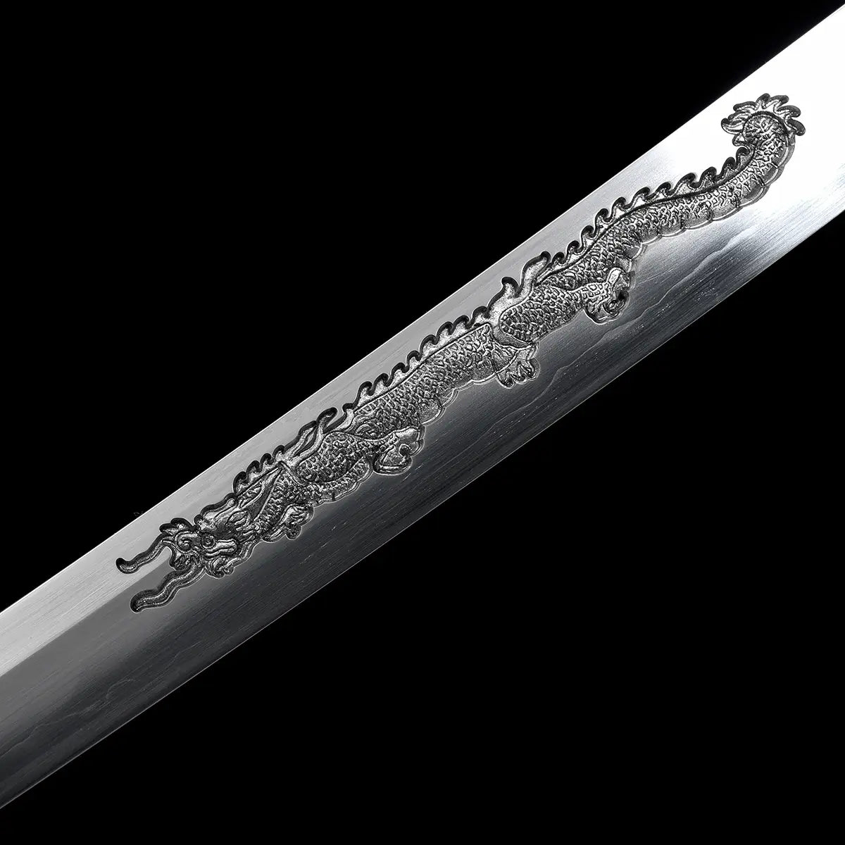 Gros plan sur la lame du katana japonais artisanal Ryū no Ken sur fond noir uniforme, montrant une gravure dragon en relief finement détaillée sur l’acier poli, écailles et courbes du dragon clairement visibles, surface de lame lisse avec reflets subtils, absence de fourreau et de support, mise au point centrée sur le travail de gravure et la finition de l’acier.
