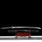 Katana japonais artisanal Ryū no Ken présenté dégainé sur un support en bois verni rouge, photographié sur fond noir avec surface inférieure gris clair, lame en acier visible avec gravure dragon sur le plat, fourreau noir laqué décoré d’un motif dragon positionné en dessous, manche noir tressé avec losanges blancs en peau de raie, garde décorative et habaki doré visibles, composition horizontale mettant en valeur la lame, le saya et le présentoir.
