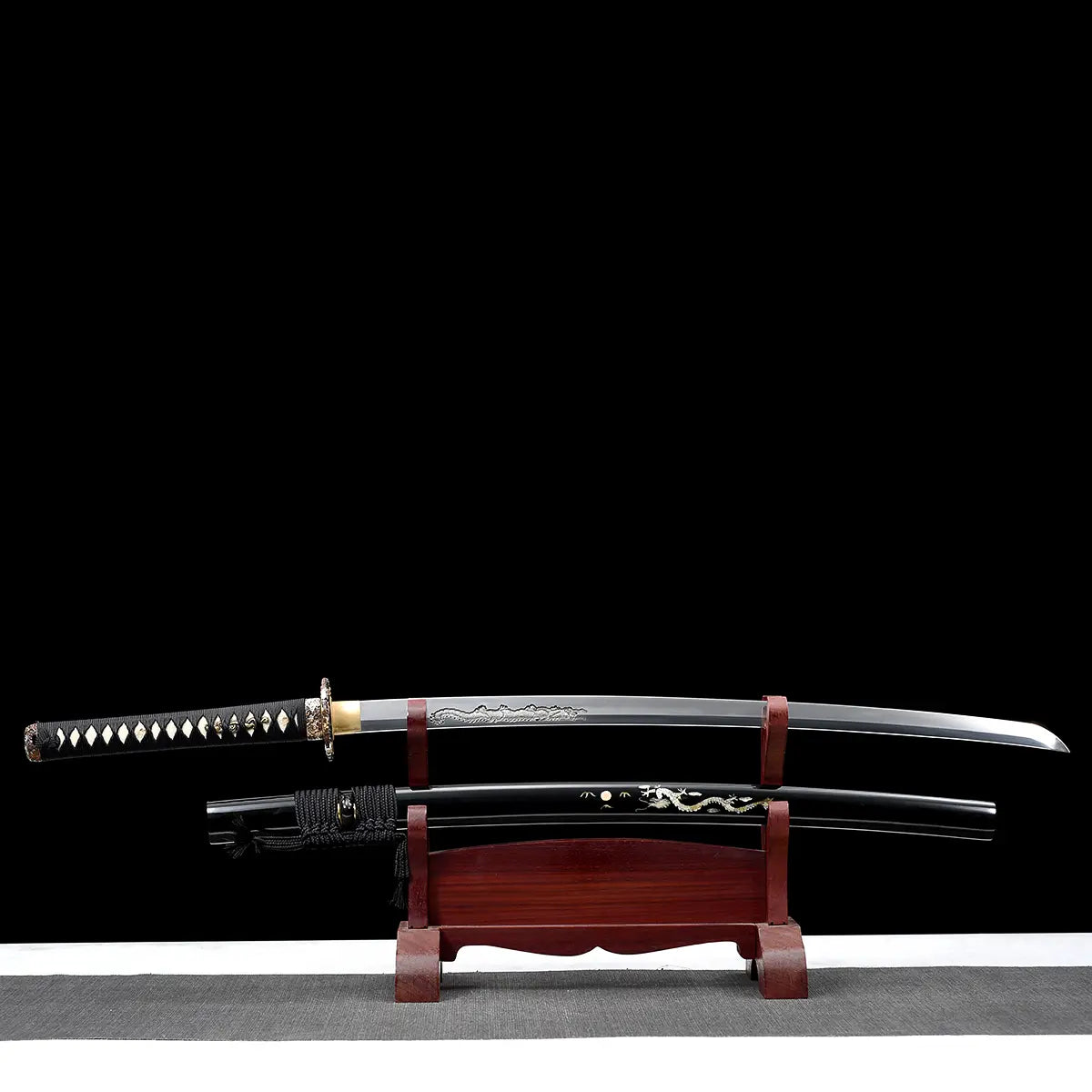 Katana japonais artisanal Ryū no Ken présenté dégainé sur un support en bois verni rouge, photographié sur fond noir avec surface inférieure gris clair, lame en acier visible avec gravure dragon sur le plat, fourreau noir laqué décoré d’un motif dragon positionné en dessous, manche noir tressé avec losanges blancs en peau de raie, garde décorative et habaki doré visibles, composition horizontale mettant en valeur la lame, le saya et le présentoir.

