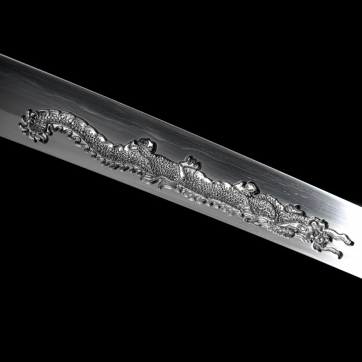 Détail du corps du dragon gravé sur la lame du katana japonais artisanal Ryū no Ken, photographié sur fond noir uni, acier poli avec reflets subtils et lignes de trempe visibles, relief du dragon finement sculpté avec écailles détaillées et ondulations marquées, absence de fourreau et de support, cadrage serré mettant en valeur la précision de la gravure et la texture de l’acier.
