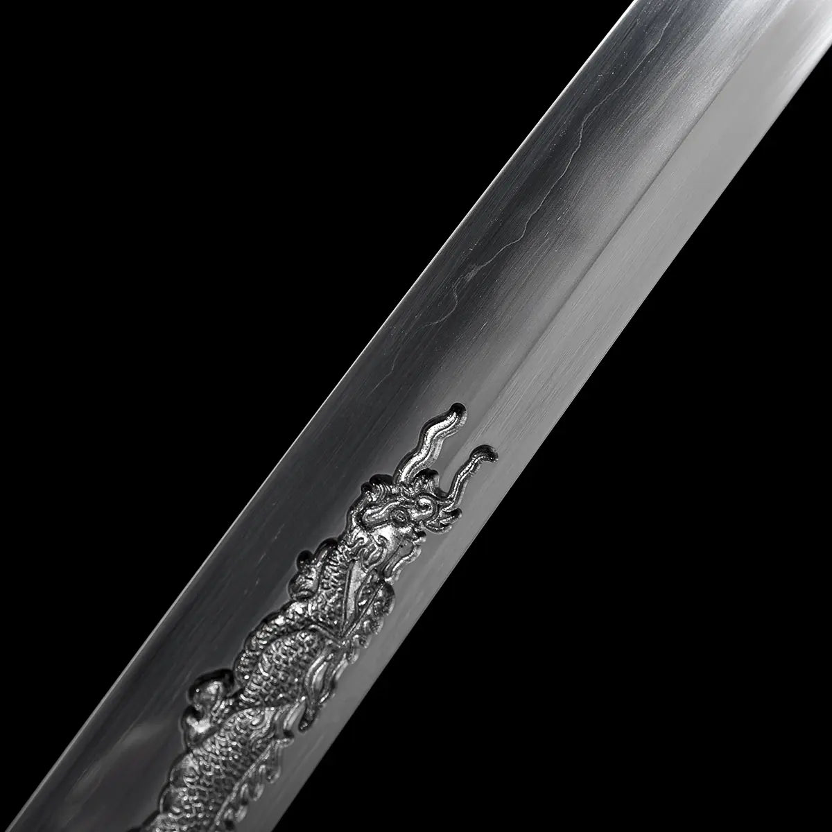 Détail rapproché de la tête du dragon gravée sur la lame du katana japonais artisanal Ryū no Ken, photographié sur fond noir profond, acier poli avec lignes de trempe visibles le long du tranchant, relief du dragon finement sculpté avec cornes et écailles apparentes, absence de fourreau et de support, mise au point centrée sur la gravure et la texture de l’acier.

