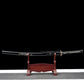 Vue complète du katana japonais artisanal Ryū no Ken présenté sur un support en bois verni rouge, photographié sur fond noir avec surface inférieure gris clair, lame rangée dans son fourreau noir laqué décoré d’un motif dragon, manche noir tressé avec losanges blancs en peau de raie, garde décorative visible, sabre posé horizontalement sur le présentoir, composition frontale mettant en valeur l’ensemble du katana et son support.
