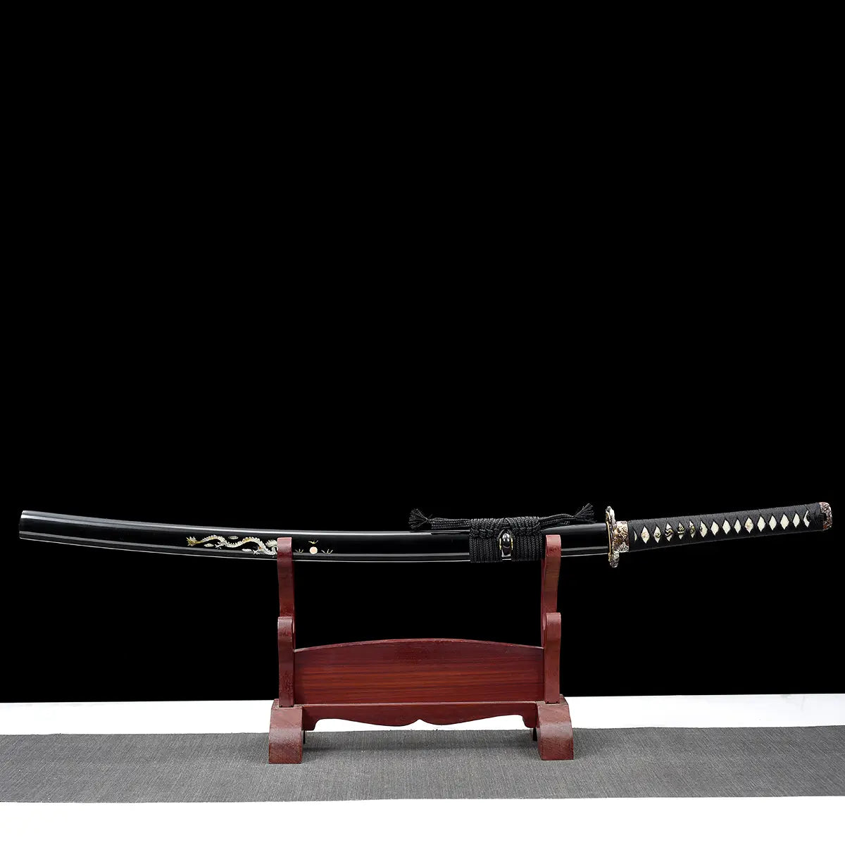 Vue complète du katana japonais artisanal Ryū no Ken présenté sur un support en bois verni rouge, photographié sur fond noir avec surface inférieure gris clair, lame rangée dans son fourreau noir laqué décoré d’un motif dragon, manche noir tressé avec losanges blancs en peau de raie, garde décorative visible, sabre posé horizontalement sur le présentoir, composition frontale mettant en valeur l’ensemble du katana et son support.
