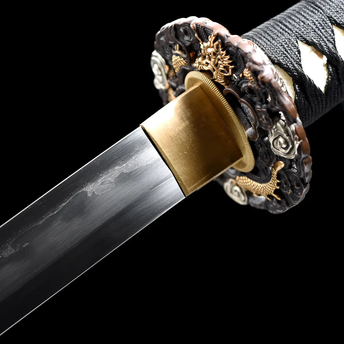 Gros plan sur la jonction entre la lame et la garde du katana japonais artisanal Ryū no Ken sur fond noir profond, lame en acier poli avec lignes de trempe visibles, habaki en laiton doré parfaitement ajusté, tsuba ronde richement sculptée avec motifs de dragon et détails dorés et argentés, manche noir tressé avec losanges blancs en peau de raie partiellement visible, absence de fourreau et de support, cadrage serré mettant en valeur la précision des ajustements et la qualité des finitions métalliques.
