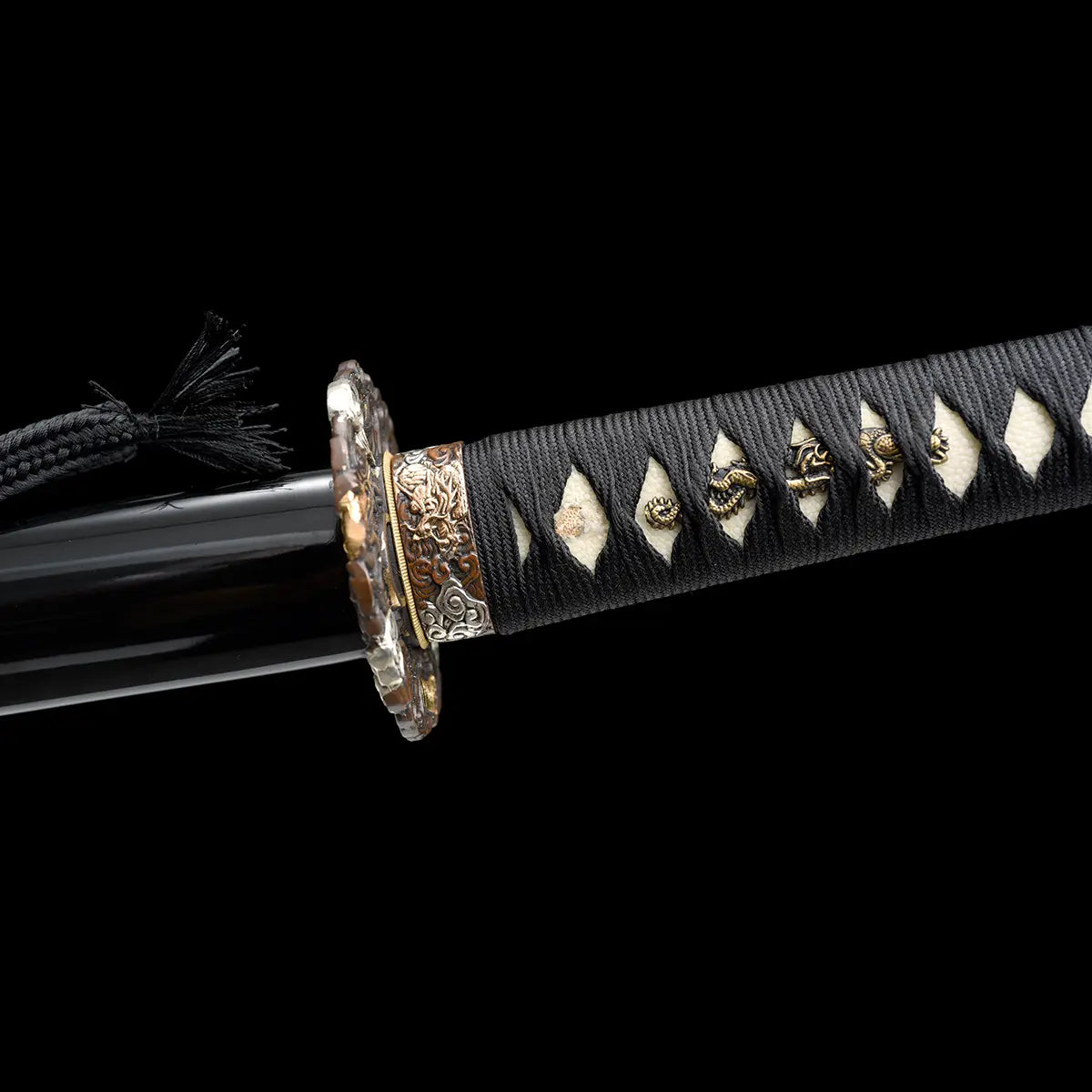 Détail du manche du katana japonais artisanal Ryū no Ken sur fond noir profond, tsuka noire tressée avec losanges blancs en peau de raie apparente et ornements métalliques décoratifs, tsuba ronde partiellement visible avec finitions sculptées, fourreau noir laqué présent à gauche avec cordon noir, absence de support, cadrage horizontal mettant en valeur la texture du tressage et la qualité des ajustements.
