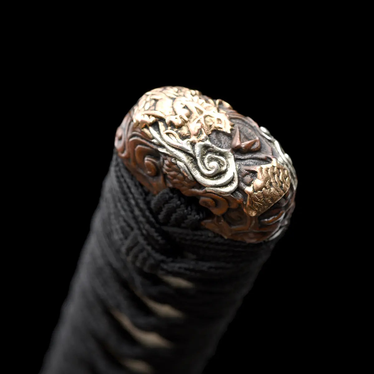 Gros plan sur l’extrémité du manche du katana japonais artisanal Ryū no Ken sur fond noir profond, kashira métallique sculptée avec motifs de dragon et détails dorés et argentés, tsuka noire tressée serrée avec texture visible, absence de lame, de fourreau et de support, cadrage centré mettant en valeur la finition du pommeau et le relief des ornements.
