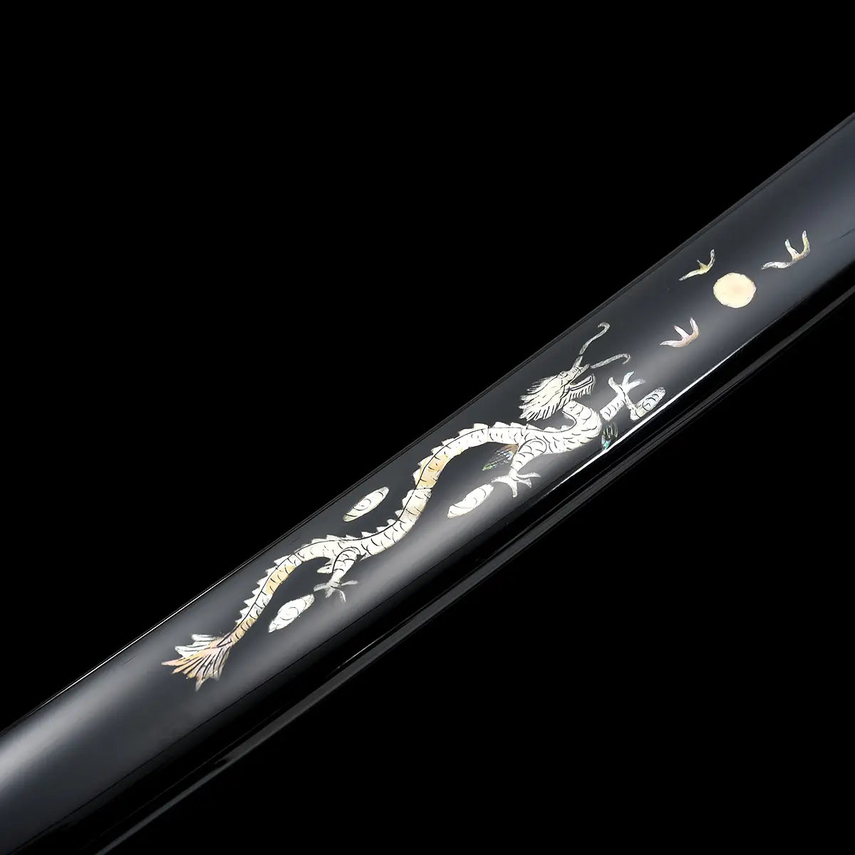 Détail du fourreau du katana japonais artisanal Ryū no Ken photographié sur fond noir uniforme, saya noir laqué brillant décoré d’un dragon incrusté clair avec éléments contrastés, motif lunaire et détails fins visibles le long du fourreau, surface lisse et réfléchissante, absence de lame apparente et de support, cadrage serré mettant en valeur le travail décoratif et la finition du saya.
