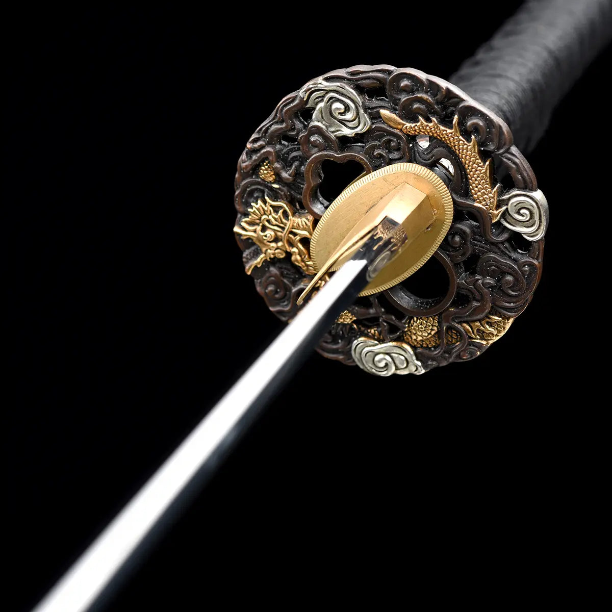 Vue frontale de la garde du katana japonais artisanal Ryū no Ken sur fond noir profond, tsuba ronde finement sculptée avec motifs de dragon et éléments décoratifs dorés et argentés, habaki en laiton doré centré, lame en acier visible dans l’axe avec reflets lumineux, manche noir tressé en arrière-plan, absence de fourreau et de support, cadrage axial mettant en valeur la symétrie et le niveau de détail de la garde.
