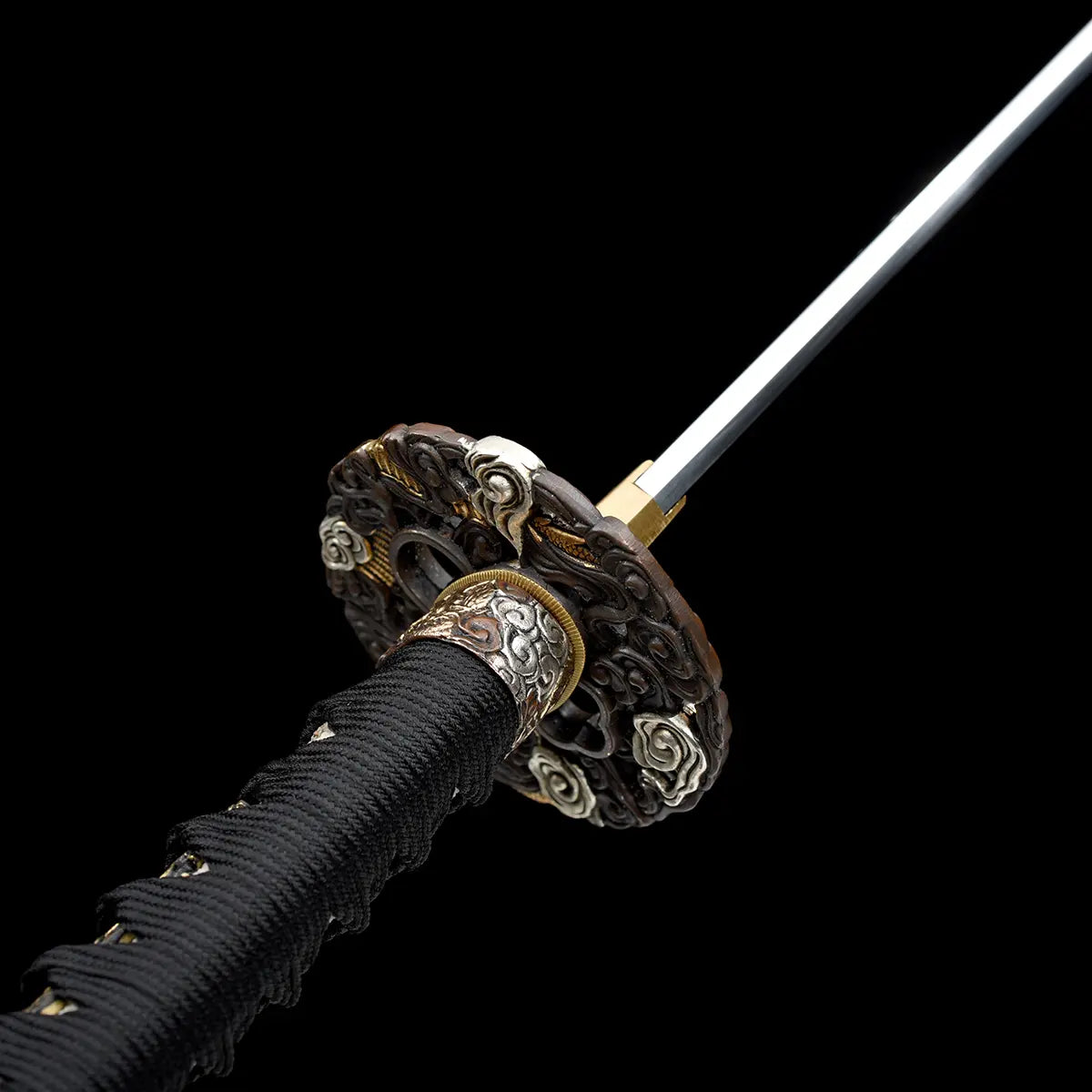 Vue en angle de la jonction lame et manche du katana japonais artisanal Ryū no Ken sur fond noir profond, lame en acier brillant orientée vers le haut, habaki en laiton doré bien ajusté, tsuba ronde richement sculptée avec motifs traditionnels et détails dorés, manche noir tressé avec losanges blancs en peau de raie visible, absence de fourreau et de support, cadrage serré mettant en valeur la garde et la finition des éléments métalliques.
