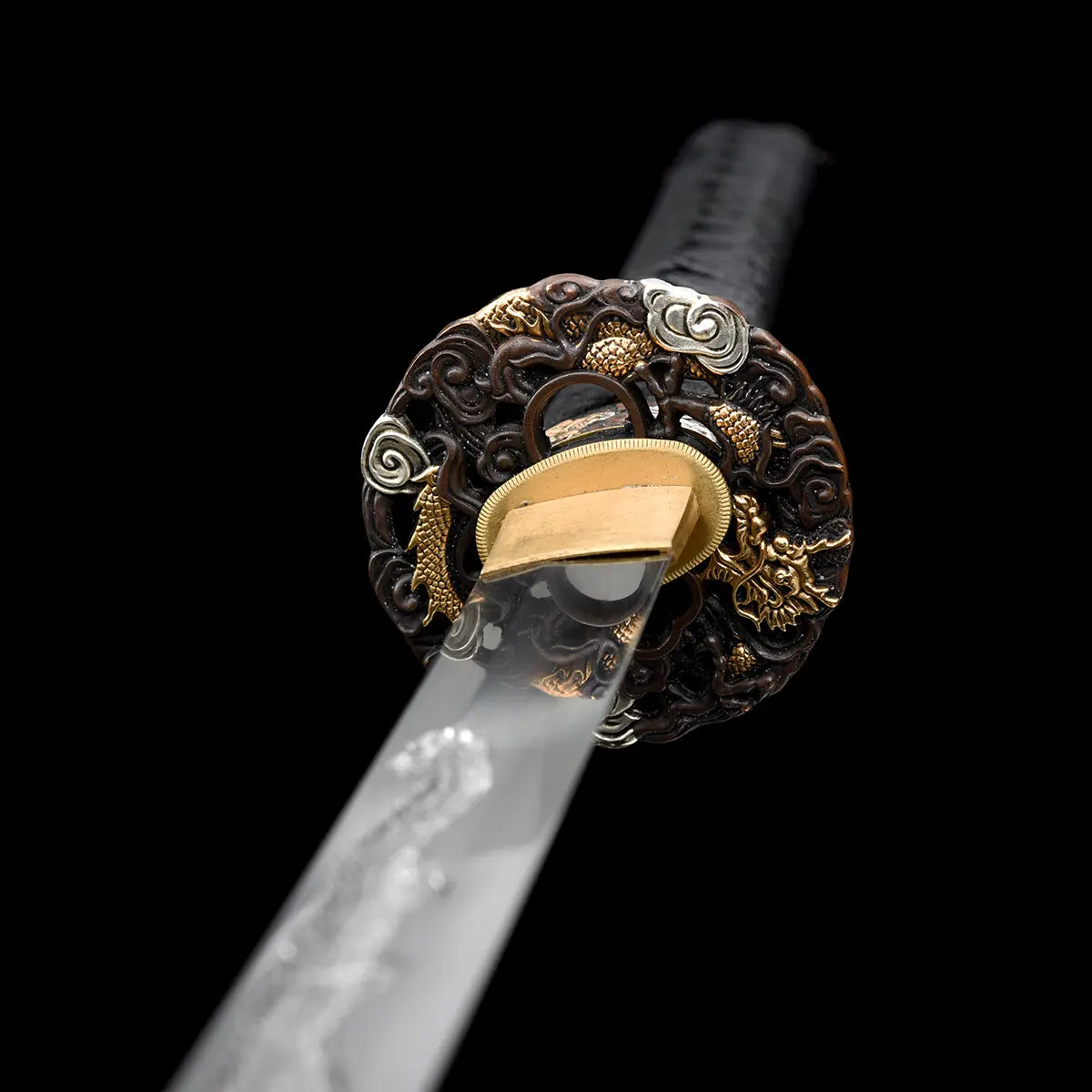 Vue arrière en angle de la garde du katana japonais artisanal Ryū no Ken sur fond noir profond, tsuba ronde sculptée avec motifs de dragon et éléments décoratifs dorés et argentés, habaki en laiton doré visible à la jonction de la lame, lame en acier partiellement floutée au premier plan, manche noir tressé en arrière-plan, absence de fourreau et de support, cadrage serré mettant en valeur la profondeur des reliefs et la finition de la garde.
