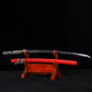 Japanese katana Sakura no Ken displayed on a wooden stand, black background, unsheathed black blade with engraved floral sakura pattern, red leather wrapped saya placed below the blade, black and white tsuka handle visible, traditional samurai sword presentation with curved blade profile
