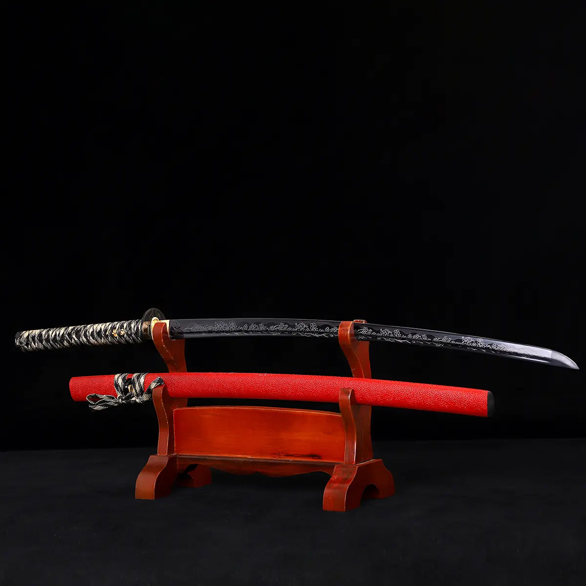 Japanese katana Sakura no Ken displayed on a wooden stand, black background, unsheathed black blade with engraved floral sakura pattern, red leather wrapped saya placed below the blade, black and white tsuka handle visible, traditional samurai sword presentation with curved blade profile
