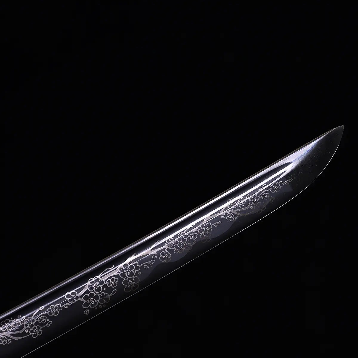 Close-up of Japanese katana Sakura no Ken blade tip, black background, unsheathed black steel blade with engraved sakura floral pattern along the edge, visible curved kissaki, decorative engraving details highlighted on the blade surface

