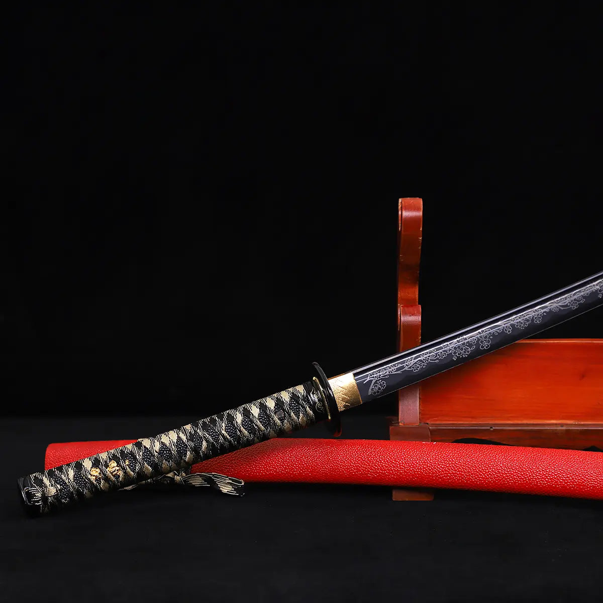 Close-up of Japanese katana Sakura no Ken blade and handle, black background, unsheathed black blade with engraved floral sakura pattern, golden habaki and dark tsuba visible, black and white tsuka wrap, red leather wrapped saya placed beneath the blade, traditi
