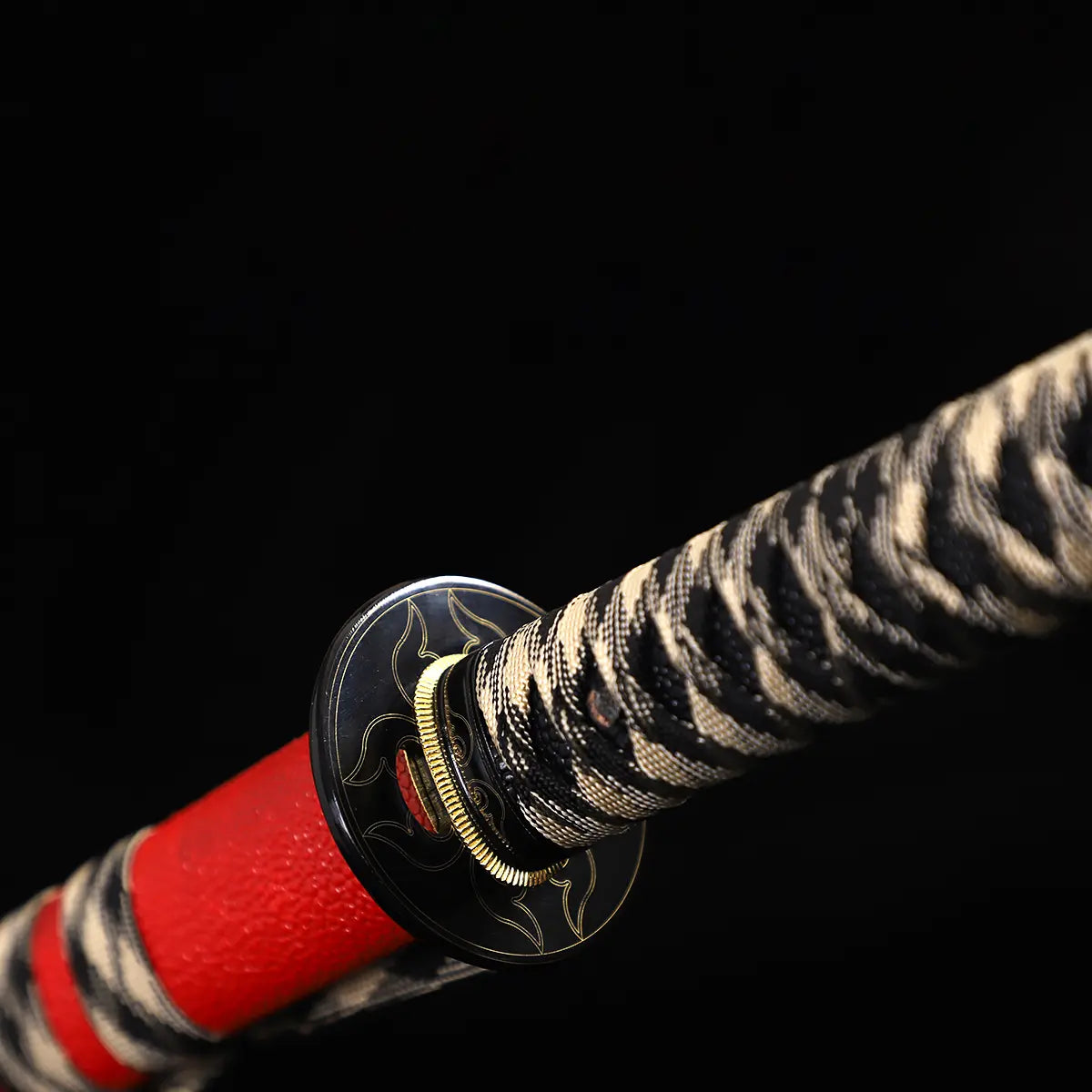 Close-up of Japanese katana Sakura no Ken handle and guard, black background, black round tsuba with engraved pattern, black and white tsuka ito wrapping over ray skin, golden habaki edge visible, red leather wrapped saya partially visible beneath the guard, traditional samurai sword mounting details
